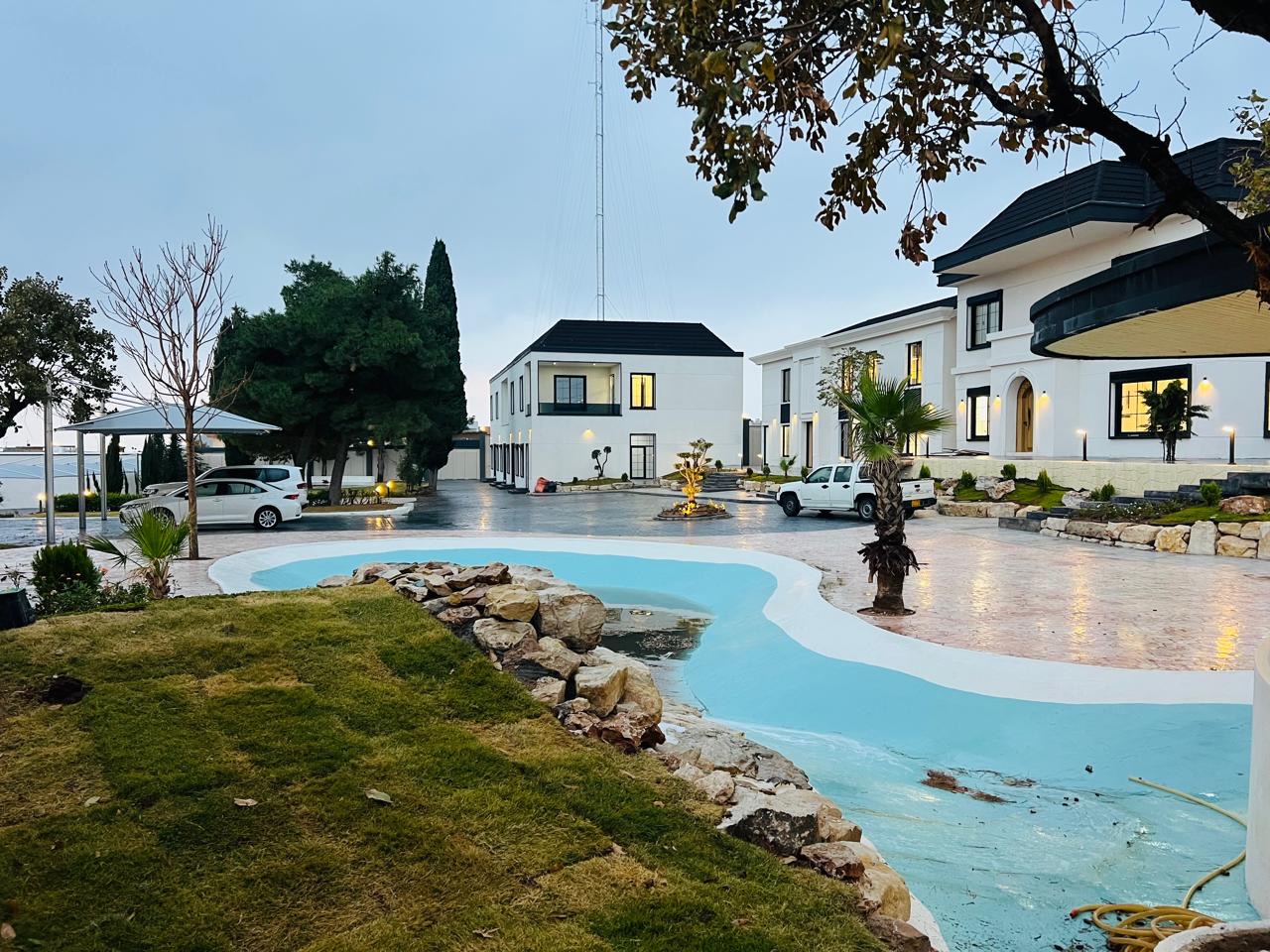 The finished compound at dusk: white buildings with lit windows beside a stone-edged pool and lawn