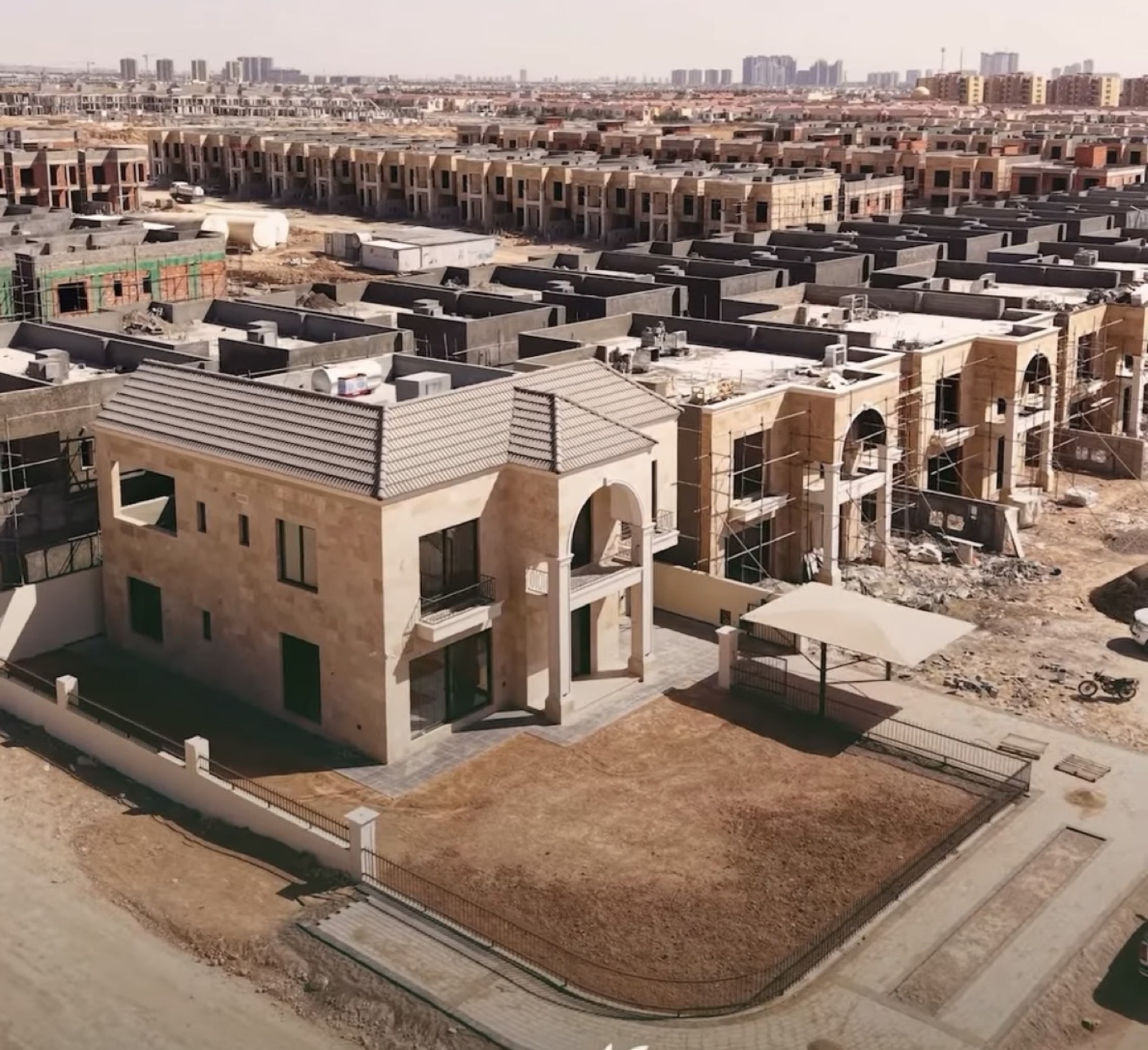 Olive Garden City: a finished stone-clad villa with a tiled roof and lawn, rows of villas under construction behind it